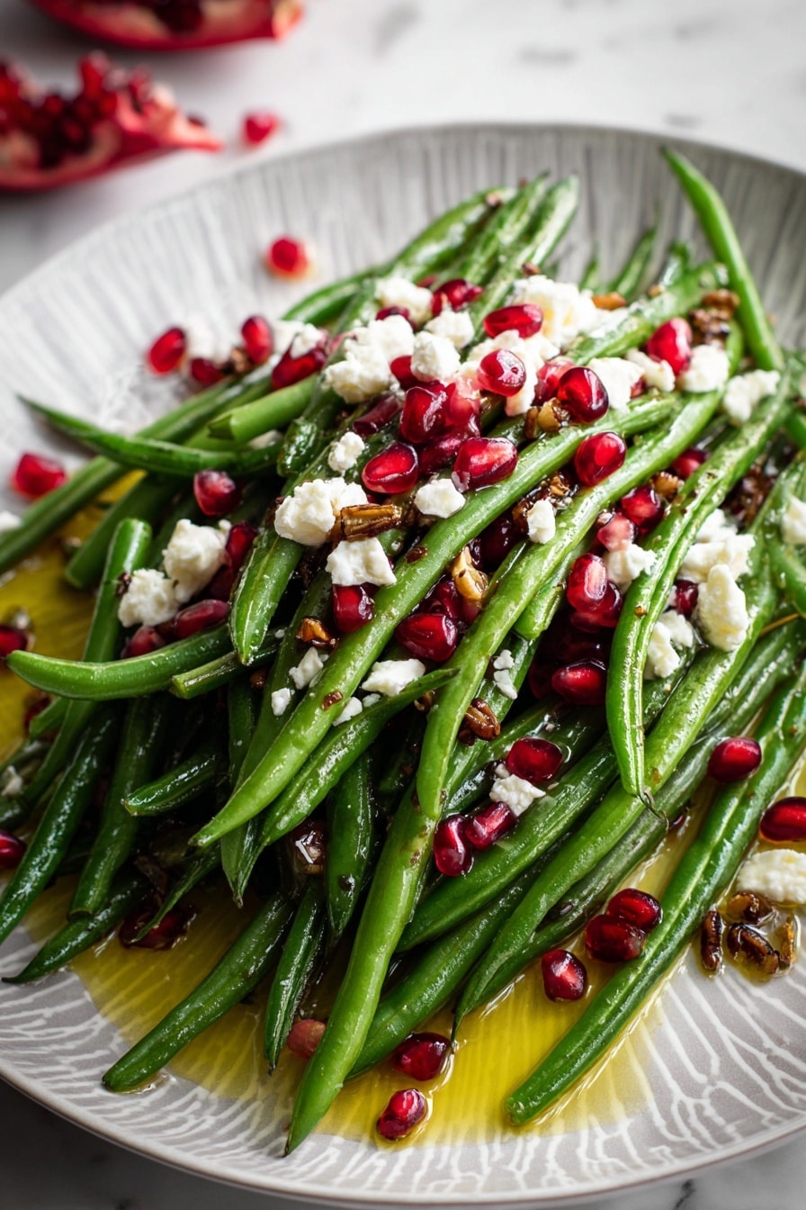 A white oval plate with a light grey pattern holds a fresh green bean salad. The bottom layer is bright green beans, long and whole, neatly piled on the plate. Scattered on top of the beans are bright red pomegranate seeds and small white crumbled cheese pieces. There are also tiny dark brown bits sprinkled over everything. The plate is placed on a white marbled surface with some pomegranate seeds around it, and a piece of pomegranate fruit is seen in the upper left corner. A bit of light dressing pools gently around the green beans. photo taken with an iphone --ar 2:3 --v 7 - Maple Mustard Green Beans with Pomegranate and Goat Cheese, healthy green bean side dish, spring green beans recipe, fruit and cheese salad, easy vegetable side dish